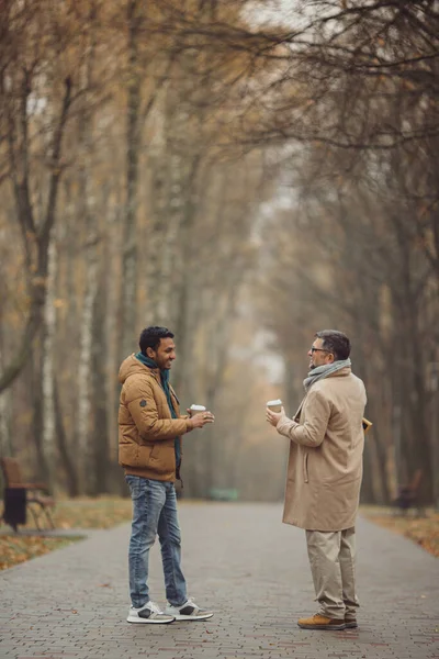 朋友们 一个老人和一个年轻人一起在秋天的公园里散步 喝咖啡 — 图库照片