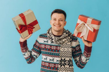 Noel süveteri ve eşarplı genç adam mavi arka planda saklanmış hediye kurdeleli bir sürü hediye kutusu tutuyor. Mutlu yıllar, kutlama konsepti..