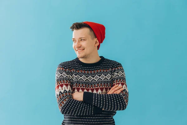 青の背景に隔離されたクリスマスセーターと赤い帽子を身に着けている魅力的な男の写真 — ストック写真