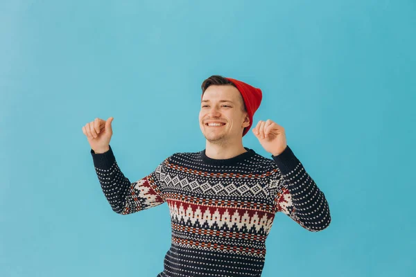 青の背景に隔離されたクリスマスセーターと赤い帽子を身に着けている魅力的な男の写真 — ストック写真
