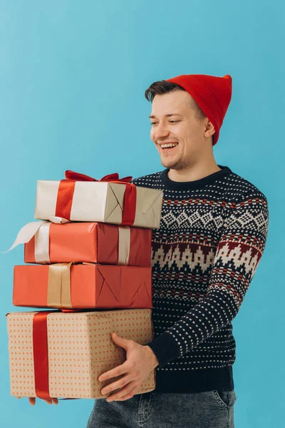 身穿圣诞毛衣头戴红帽子的年轻人 拿着许多礼品盒 上面有蓝色背景的彩带蝴蝶结 新年快乐 庆祝理念 — 图库照片