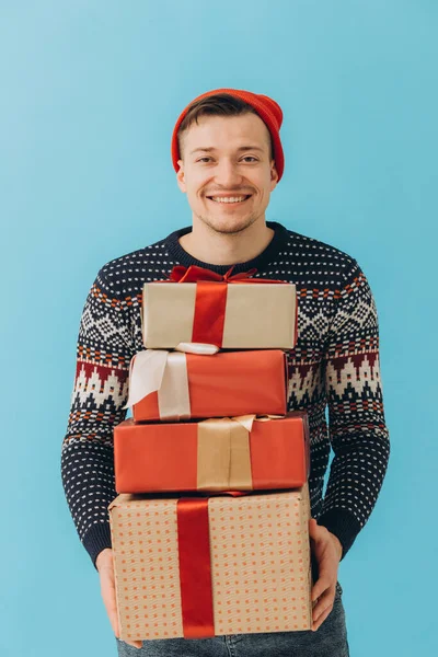身穿圣诞毛衣头戴红帽子的年轻人 拿着许多礼品盒 上面有蓝色背景的彩带蝴蝶结 新年快乐 庆祝理念 — 图库照片