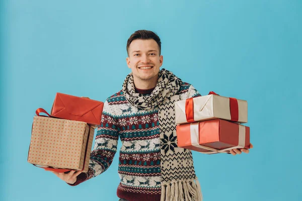 クリスマスのセーターやスカーフの若い男は ブルーの背景に孤立ギフトリボン弓と多くのギフトボックスを保持します 新年おめでとうお祝いのコンセプト — ストック写真