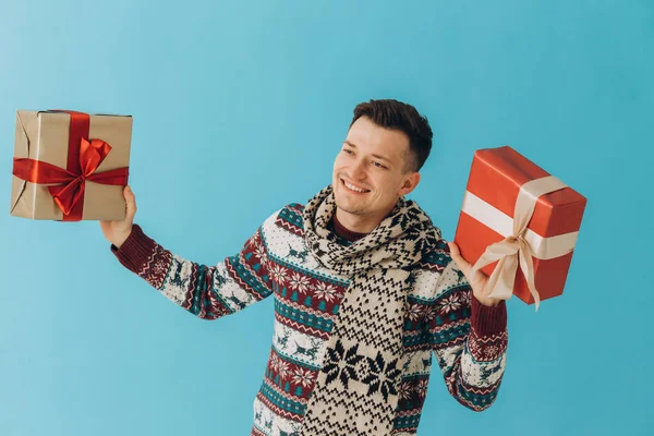 クリスマスのセーターやスカーフの若い男は ブルーの背景に孤立ギフトリボン弓と多くのギフトボックスを保持します 新年おめでとうお祝いのコンセプト — ストック写真