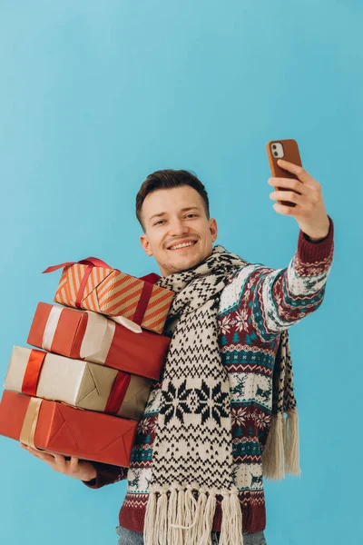クリスマスのセーターやスカーフの若い男は 多くのギフトボックスを保持し 青の背景に隔離された自撮りを取ります 新年おめでとうお祝いのコンセプト — ストック写真