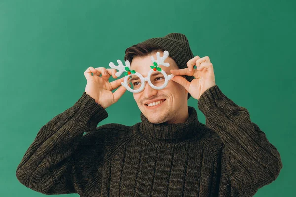 一个穿着保暖毛衣 头戴帽子 戴着狂欢节眼镜 被绿色背景隔离的年轻人 — 图库照片