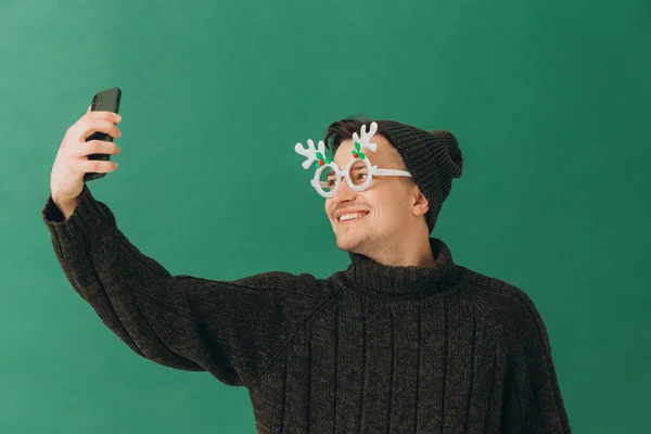 緑の背景に隔離された彼の手の中に暖かいセーター カーニバルの眼鏡 電話で若い男 — ストック写真