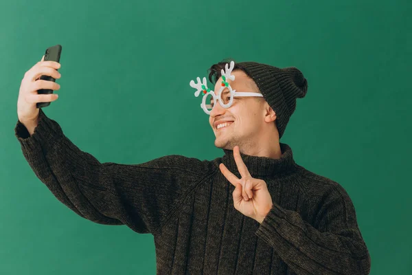 緑の背景に隔離された彼の手の中に暖かいセーター カーニバルの眼鏡 電話で若い男 — ストック写真