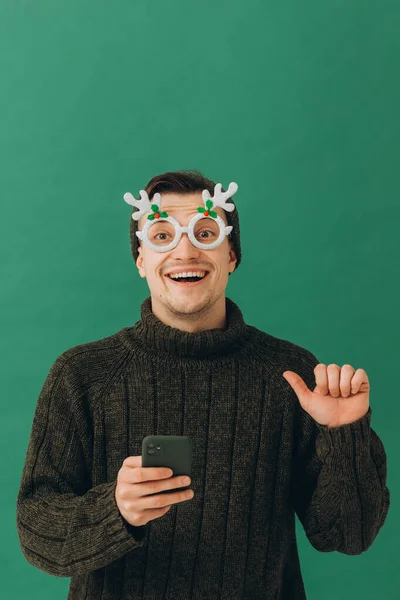 一个穿着保暖毛衣 戴着狂欢节眼镜 手里拿着手机的年轻人 被绿色的背景隔开了 — 图库照片
