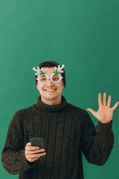 一个穿着保暖毛衣 戴着狂欢节眼镜 手里拿着手机的年轻人 被绿色的背景隔开了 — 图库照片