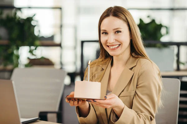 Happy businesswoman holding cake with lightened candle in the office and looking at camera.