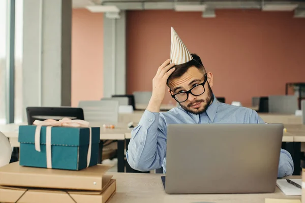 After birthday party. Bearded office worker in cone hat sitting at workplace with laptop and gift with festive ribbon.