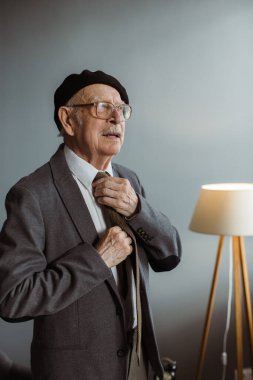 Evdeki oturma odasında duran gri saçlı, gözlüklü yaşlı bir adamın portresi..