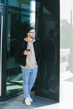 Online communication. A young man in a hat and glasses with a smartphone in his hand on the street. A young bearded guy on an urban background is chatting on a mobile phone.