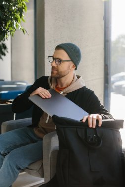 Young handsome freelancer guy working with laptop in office space. A man in glasses and a hat is holding a laptop while sitting in a chair in the hall.