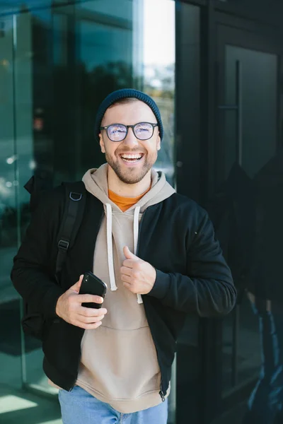 Positive young guy holding smartphone in his hands, looking at camera standing on street. Bearded man in eyeglass wears casual clothes. Cell phone usage concept