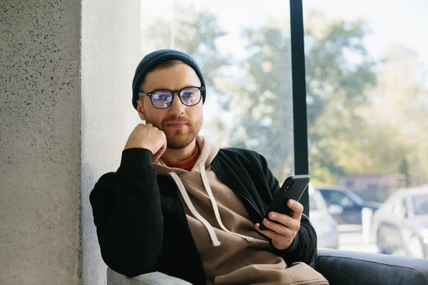 A young handsome guy with a phone in his hands is waiting for a job interview in an office space. A man in glasses and a cap is holding a phone while sitting in a chair in the hall.