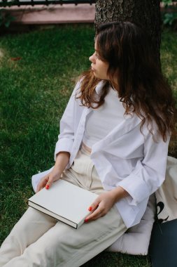 Dergi ya da kitap tasviri. Kız kahve dükkanının bahçesindeki çimlerde rahatlıyor, kitap okuyor..