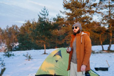 Çadırın yanında bir fincan çay ya da kahveyle duran bir turist, kış ormanında günbatımının tadını çıkarır..