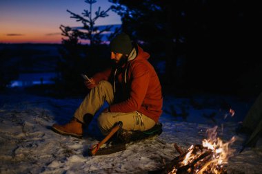 Yürüyüşçünün biri, geceleri dağlarda kamp ateşinde kendini ısıtırken telefonunu kontrol eder..
