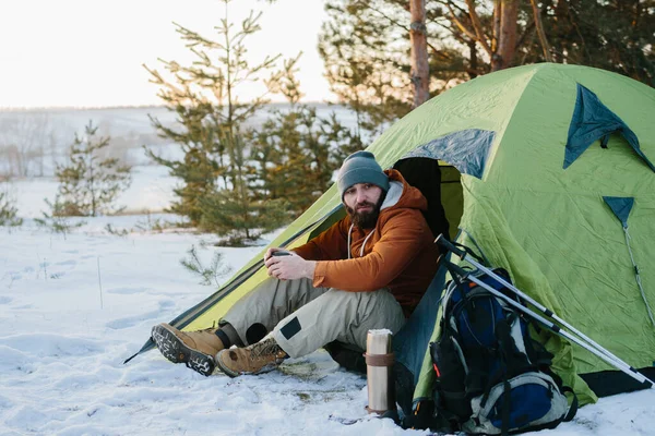 一个留着胡子的年轻人躺在靠近帐篷的冬山里 一个头戴大胡子 身穿保暖夹克的男人 徒步旅行后喝热茶或咖啡 生活方式 — 图库照片