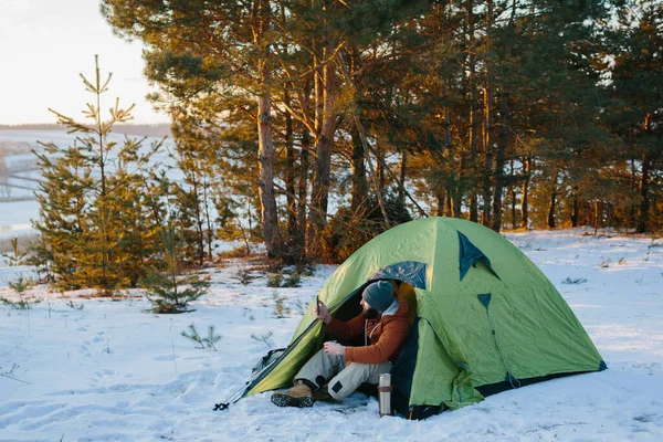 年轻的留着胡子的男人笑着 喝咖啡 在靠近帐篷的冬山里自作主张 男性旅行者带着胡子 头戴礼帽 身穿保暖夹克 徒步旅行后在手机上自画像 生活方式 — 图库照片