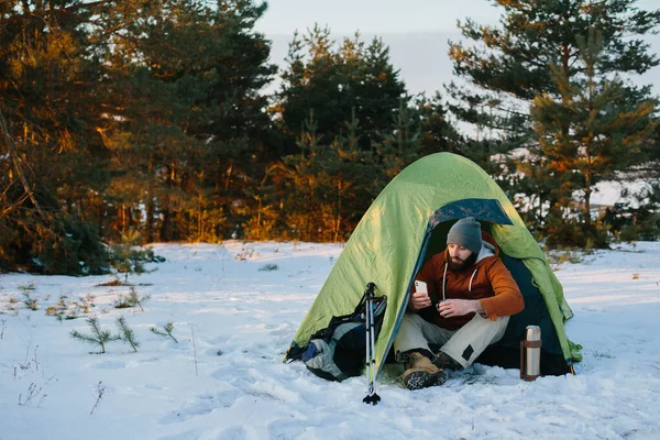 一位疲惫的游客在雪地森林的帐篷里坐着艰难的攀登之后休息了下来 男人用他的电话来传递信息或检查他的Gps — 图库照片