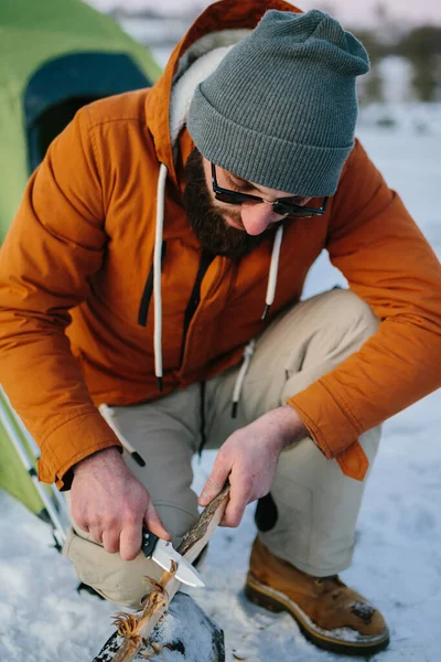 stock image A man prepares wood wool with a knife for starting a fire in a winter forest at sunset. Winter survival concept.