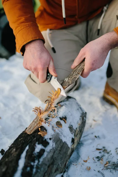 男は日没時に冬の森の中で火を起こすためのナイフで木製のウールを準備します 冬のサバイバルコンセプト — ストック写真