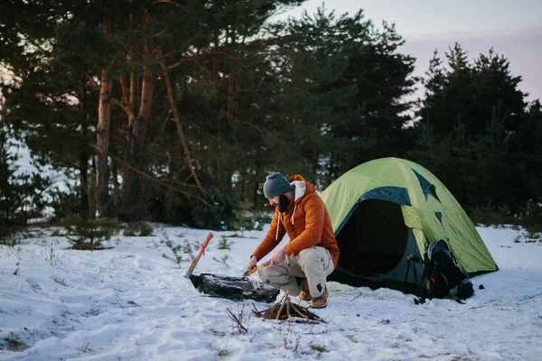 テントの近くに座っている男は 日没時に冬の森の中で火を起こすためのナイフで木製のウールを準備します 冬のサバイバルコンセプト — ストック写真
