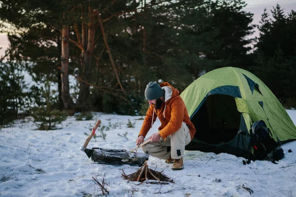 テントの近くに座っている男は 日没時に冬の森の中で火を起こすためのナイフで木製のウールを準備します 冬のサバイバルコンセプト — ストック写真