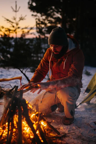 A man sits near a campfire, warming himself by the fire in a winter forest at sunset.