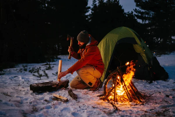 A tourist chops wood for a bonfire in a winter forest at night.