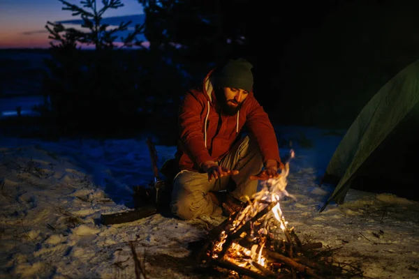 A man sits near a campfire, warming himself by the fire in a winter forest at sunset.