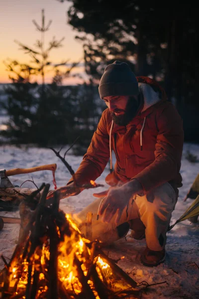 男はキャンプファイヤーの近くに座って 日没時に冬の森の中の火によって自分自身を暖める — ストック写真