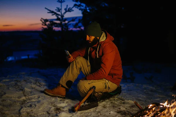一个徒步旅行者在山上的篝火边取暖 一边检查他的手机 — 图库照片