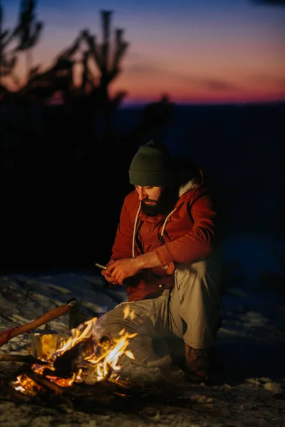 山の中で夜のキャンプファイヤーで体を暖めながら携帯電話をチェックするハイカー — ストック写真