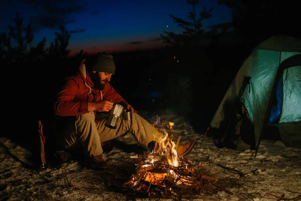 一个留着胡子的年轻人一边在篝火上煮美味的咖啡 一边在冬日的森林里欣赏日落 野外生存和远足的概念 — 图库照片