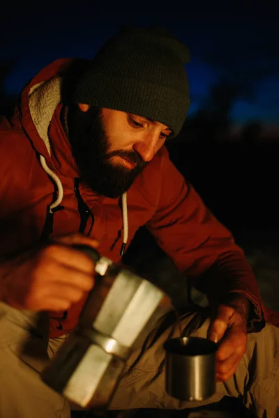冬の森で夕日を楽しみながら 髭を生やした若い男がたき火でおいしいコーヒーを作っています 野生での生存とハイキングの概念 — ストック写真
