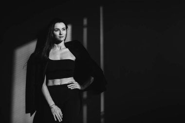Güzel bir esmer siyah ceketli gri bir duvarın arkasında poz veriyor. Üstü de camdan yansıyor..