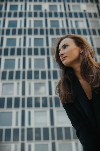 Fashion street style portrait of a beautiful girl in a black suit. A beautiful brunette poses against the background of high-rise buildings in the city.