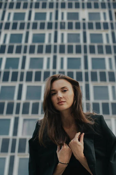 Fashion street style portrait of a beautiful girl in a black suit. A beautiful brunette poses against the background of high-rise buildings in the city.