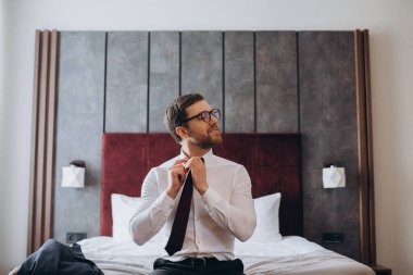 Young businessman taking off his tie while sitting on a bed in a hotel room. clipart