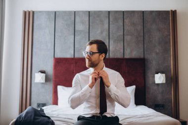 Genç işadamı otel odasında bir yatakta otururken kravatını çıkarıyor..