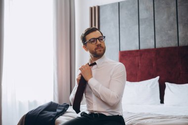 Genç işadamı otel odasında bir yatakta otururken kravatını çıkarıyor..