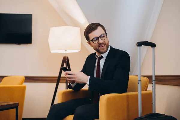 stock image A businessman checks his phone while sitting in a comfortable chair in a hotel room.