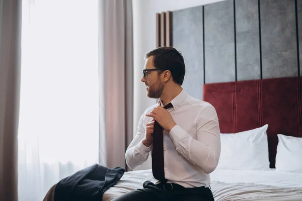 Stock image Young businessman taking off his tie while sitting on a bed in a hotel room.