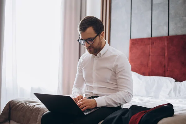 コンピュータを扱う若いビジネスマンがホテルの部屋のベッドに座っている — ストック写真