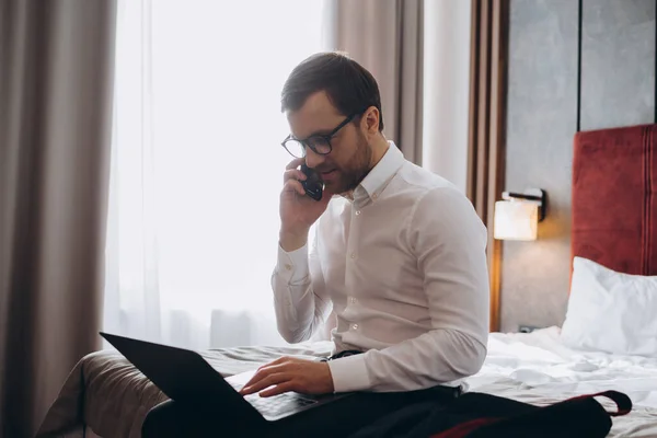 ビジネス旅行中にホテルのベッドに座ってノートパソコンを使用している間に電話をかけるハンサムなビジネスマン — ストック写真