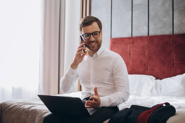 ビジネス旅行中にホテルのベッドに座ってノートパソコンを使用している間に電話をかけるハンサムなビジネスマン — ストック写真